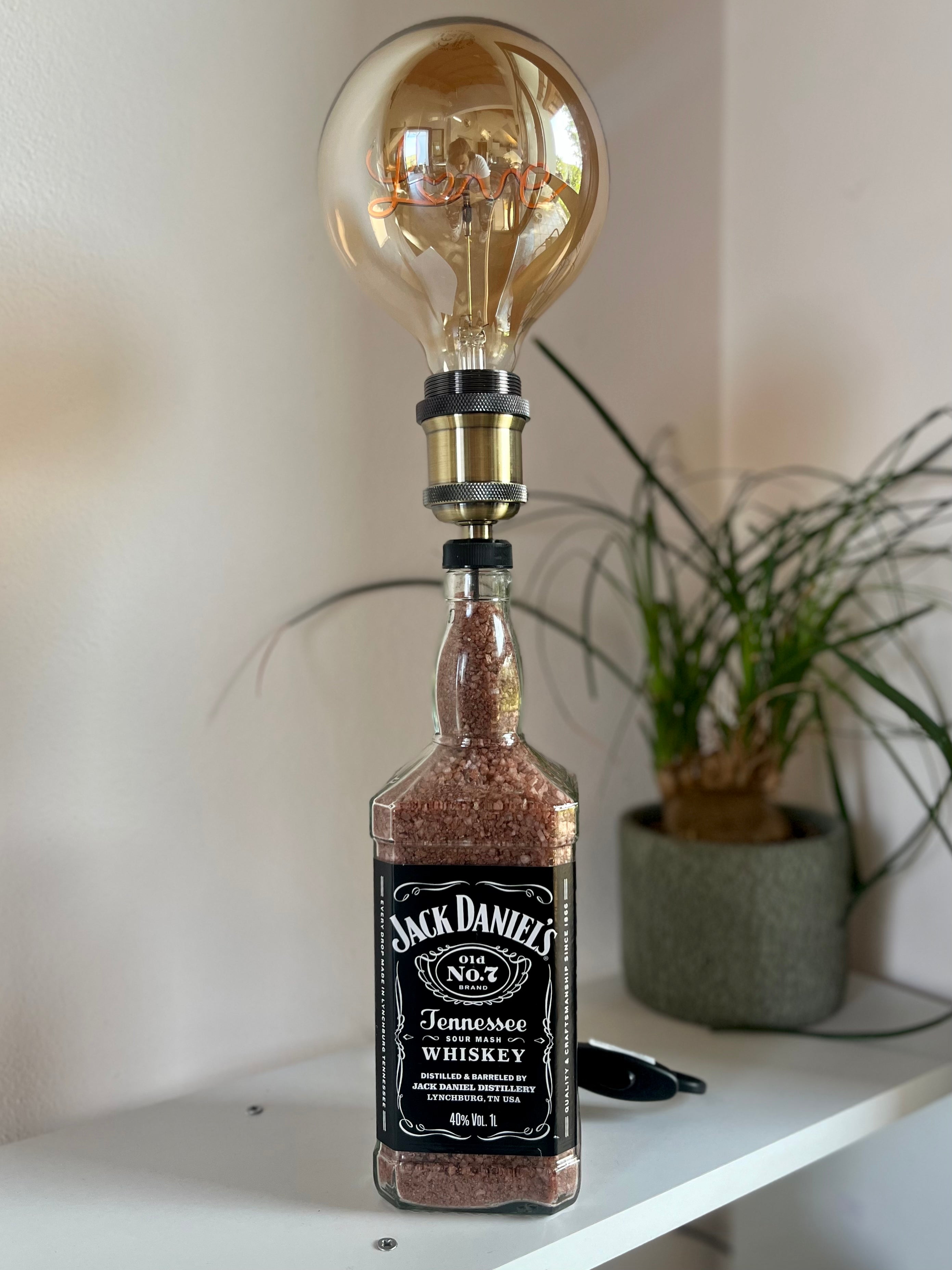 Lampada artigianale da tavolo realizzata con bottiglia Jack Daniel’s, lampadina vintage “Love” accesa, posizionata su mobile di design chiaro