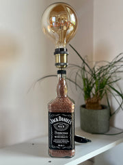 Lampada artigianale da tavolo realizzata con bottiglia Jack Daniel’s, lampadina vintage “Love” accesa, posizionata su mobile di design chiaro