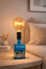 Lampada da tavolo artigianale realizzata con bottiglia di gin Portofino riciclata, con lampadina vintage a luce calda “Dreams”, posizionata su comodino in legno in una camera da letto dai toni neutri e atmosfera accogliente.