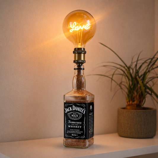 Lampada artigianale da tavolo realizzata con bottiglia Jack Daniel’s, lampadina vintage “Love” accesa, posizionata su mobile di design chiaro in ambiente domestico moderno e minimalista, luce calda e atmosfera accogliente