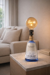 Lampada di design realizzata con bottiglia Gin Mare e lampadina vintage luminosa, su tavolino in marmo in soggiorno elegante con divano beige e luce soft
