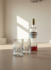 Set bicchieri in vetro riciclato da bottiglie Campari Bitter con texture ispirata al Duomo di Milano, su tavola apparecchiata moderna con luce naturale
