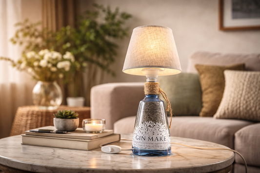 Lampada artigianale in bottiglia di gin riciclata con paralume beige, luce calda, su tavolino in marmo in soggiorno moderno con libri, pianta e candela