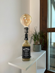 Lampada originale da tavolo realizzata con bottiglia Jack Daniel’s, lampadina vintage “Love”, posizionata su mobile di design chiaro in ambiente domestico moderno e minimalista, luce calda e atmosfera accogliente