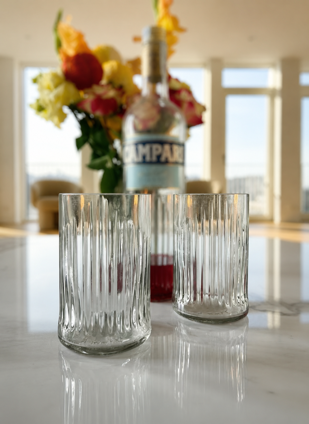 Bicchieri in vetro riciclato da bottiglie Bitter con lavorazione a rilievo ispirata al Duomo di Milano, disposti su tavola apparecchiata elegante in stile minimal, luce naturale calda e atmosfera raffinata