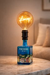 Lampada in bottiglia di gin Portofino riciclata con luce calda, posizionata su tavolino in legno accanto a pianta decorativa in ambiente domestico luminoso e naturale