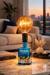 Lampada di design in bottiglia di gin Portofino con lampadina vintage accesa, su superficie neutra con sfondo minimal per evidenziare materiali, luce e dettagli artigianali