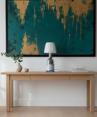 Lampada da tavolo realizzata con bottiglia di gin riciclata, con paralume in tessuto beige e corda decorativa, appoggiata su un tavolino in legno, quadro su parete